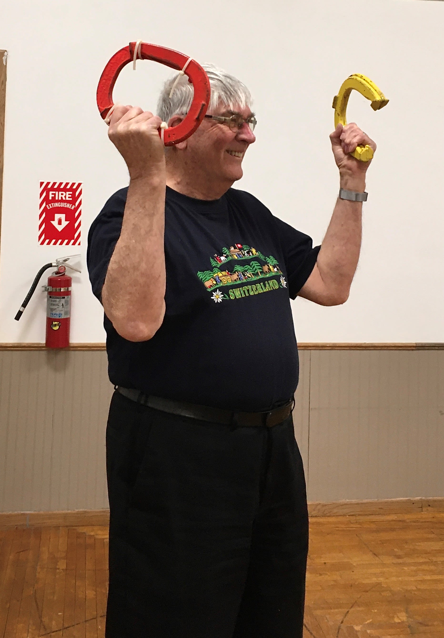 A smiling older man in a black t-shirt lifts horseshoes above his head in a fitness class. 
