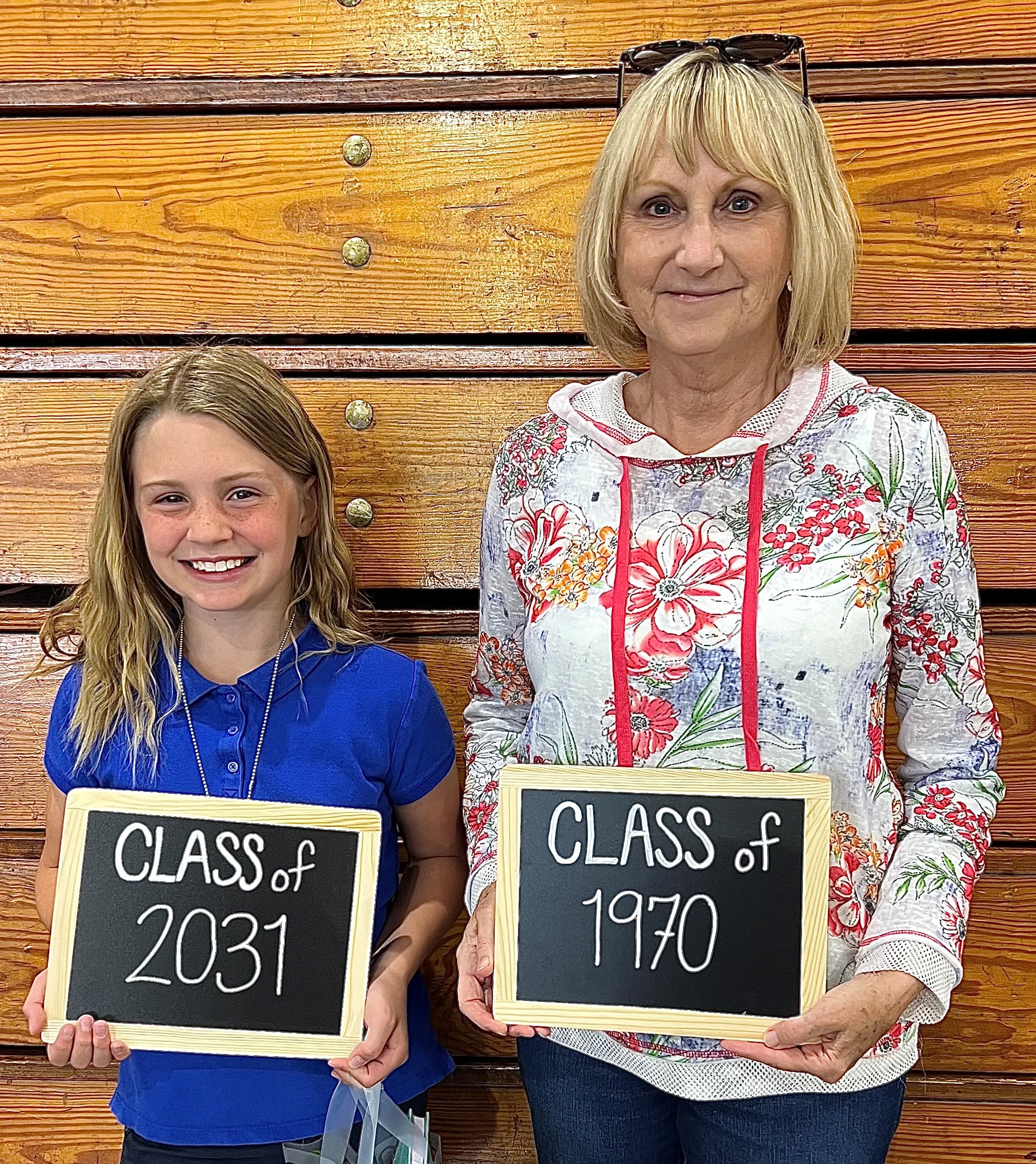 A smiling little girl stands with an older woman. The girl holds a chalkboard reading "Class of 2031" while the woman's reads "Class of 1970." They are pen pals. 