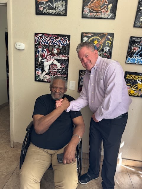 A smiling man in a wheelchair grips hands with a standing man in an Oxford shirt. Both are smiling.