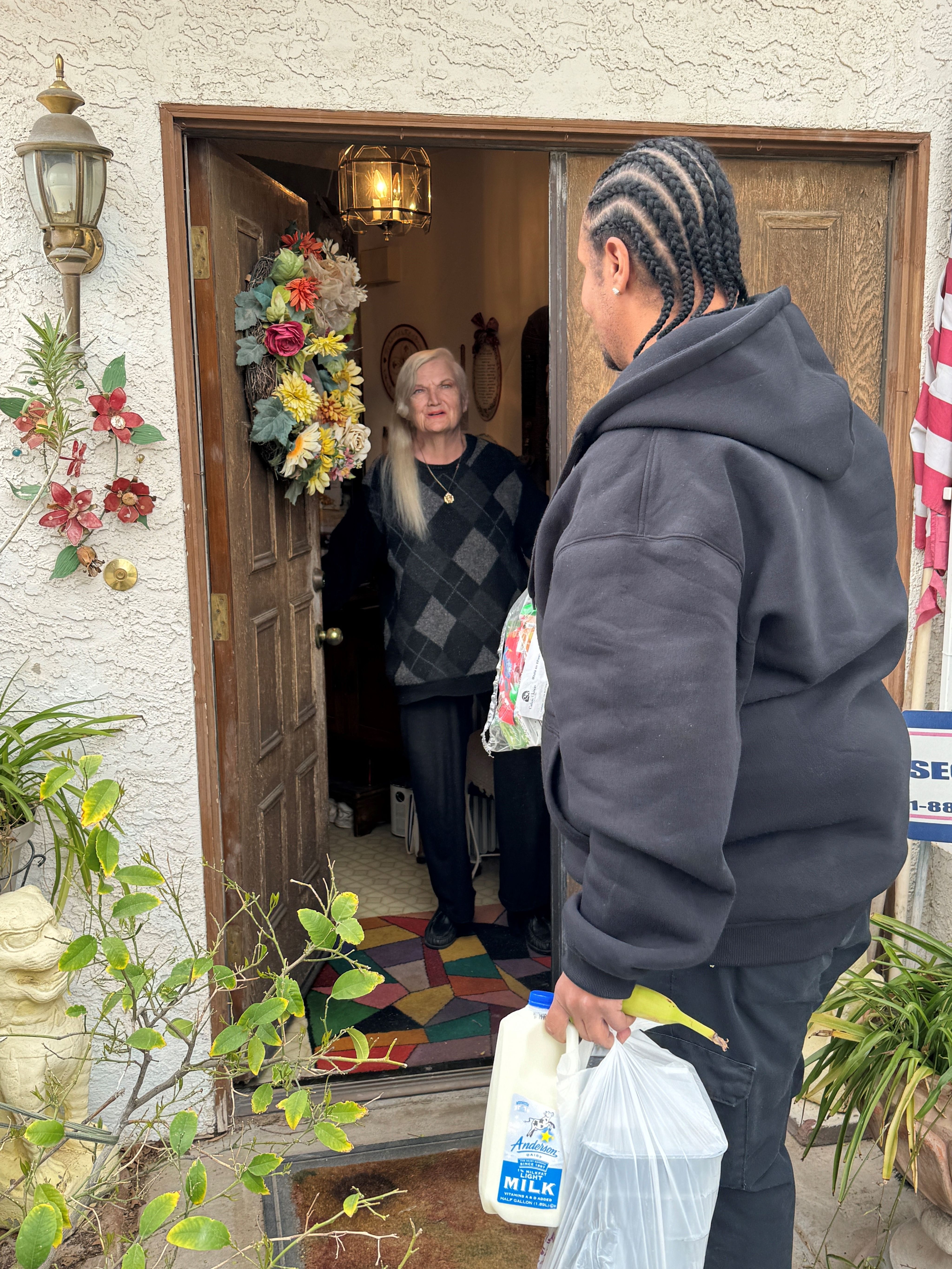 An older woman in an argyle sweater stands in her doorway to receive a delivery of milk and other groceries from a man in a hooded sweatshirt. 